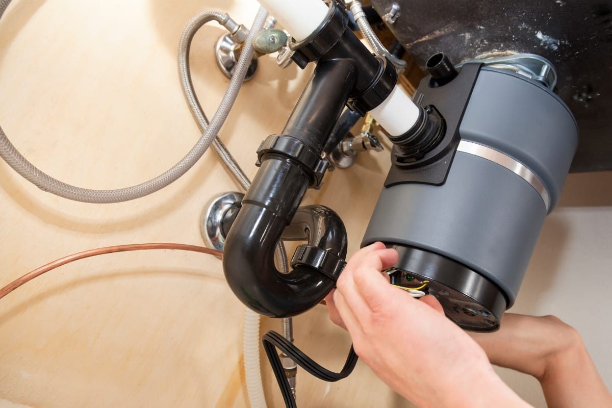 A person is expertly installing a garbage disposal unit under the kitchen sink, integrating it seamlessly with other kitchen appliances for a more efficient and modern culinary space.