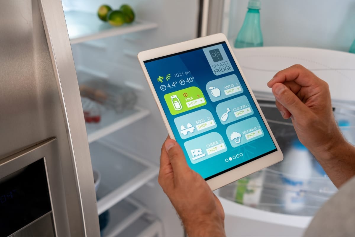 A person uses a tablet to check the smart fridge's inventory, showcasing items like milk, apples, and chicken with convenient shopping options. The open fridge door reveals some kitchen appliances discreetly nestled beside fresh groceries.