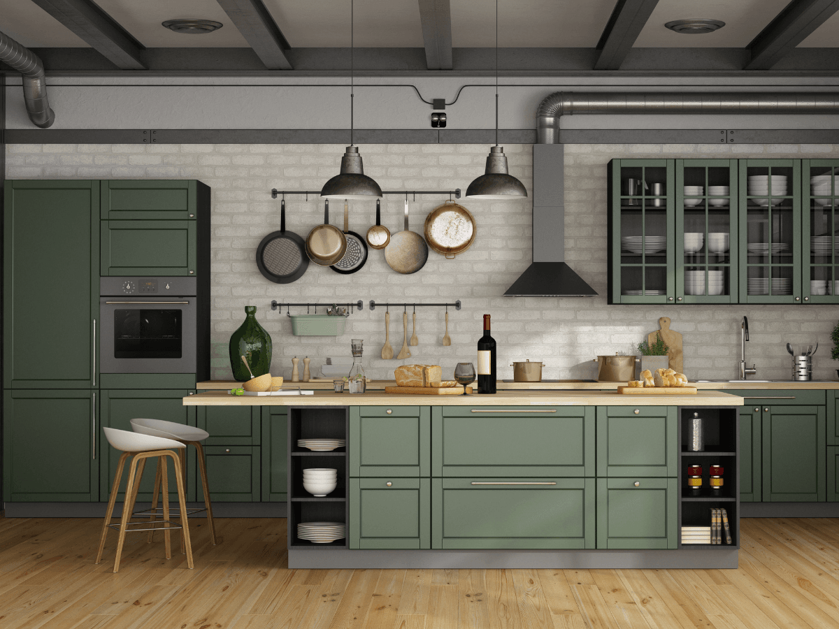 A modern kitchen with green cabinets, a central island, hanging pots, and cutting boards exudes a vintage style. A wine bottle and stools add charm, while an exposed brick wall and industrial lighting complete the look.