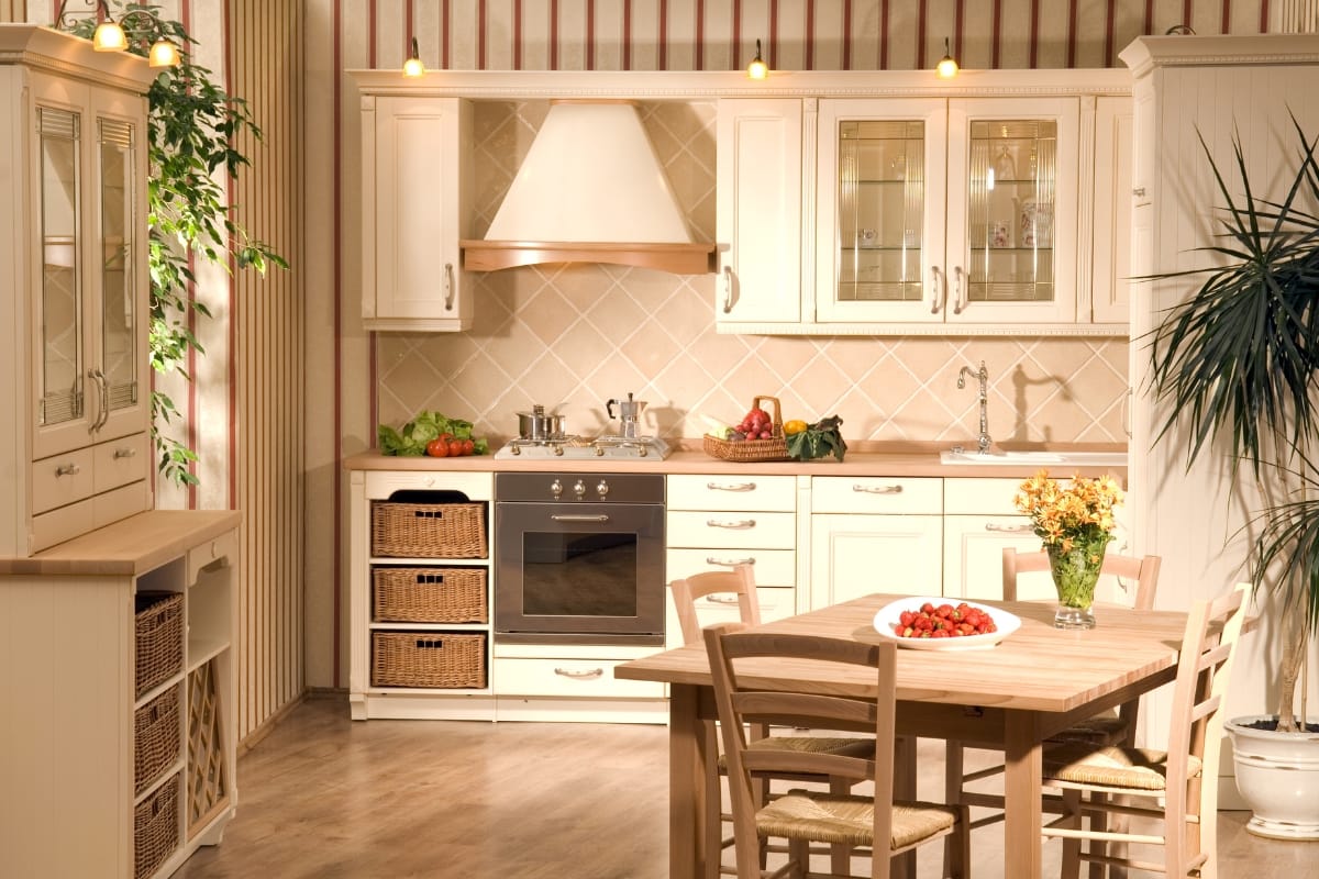 A cozy kitchen boasting vintage style with wooden cabinets, a table with chairs, and a potted plant. A white stove and range hood are surrounded by assorted vegetables and fruit on the counter. Warm lighting from ceiling fixtures enhances its nostalgic charm.