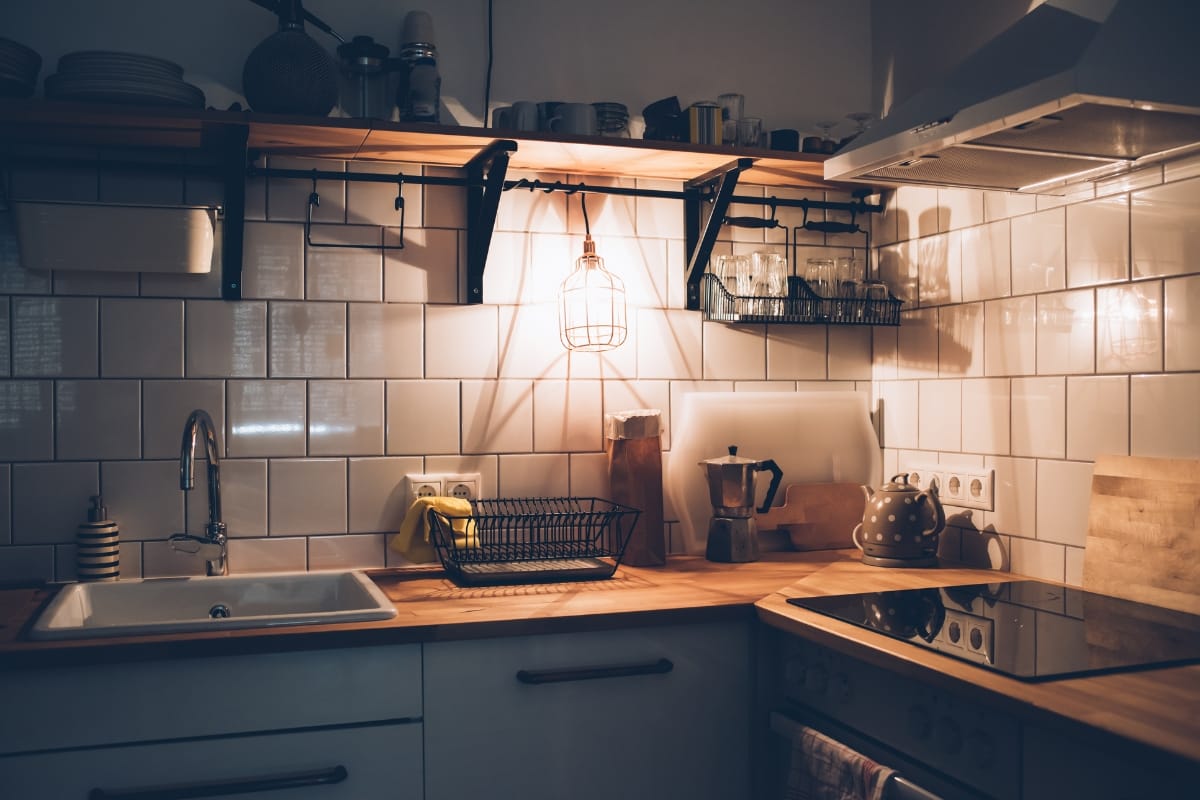 A cozy kitchen with white tiled walls and wooden countertops embodies a vintage style. The sink, kettle, stovetop, and dish rack blend seamlessly as warm light illuminates the inviting space.