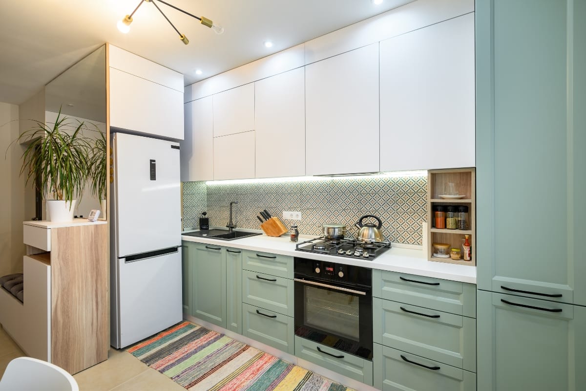 A modern kitchen with mint green cabinets evokes a hint of vintage style, complemented by a white fridge, stove, oven, and a colorful rug. A potted plant sits near the refrigerator. Bright lighting and a patterned backsplash complete the charming look.