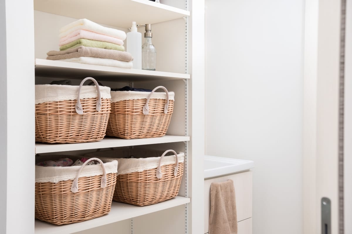 Closet with four wicker baskets on shelves, neatly folded towels, and a soap dispenser. A white sink with a beige towel is visible in the background, showcasing ways of maximizing bathroom space efficiently.