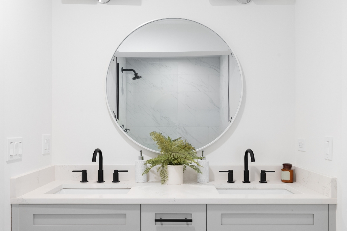 Elegant Bathroom Vanities in Roswell: Choosing the Right Style for Your Space 1 An elegant bathroom vanity with two sinks, black fixtures, a round mirror, a potted fern, and a small brown container sits atop a crisp white countertop.