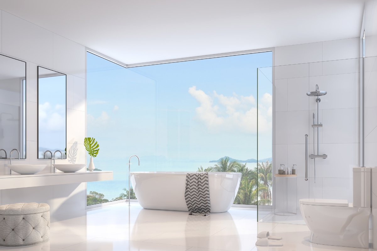 Elegant Bathroom Vanities in Roswell: Choosing the Right Style for Your Space 3 Modern white bathroom with elegant bathroom vanities, a freestanding bathtub, large windows showing an ocean view, double sinks, glass shower, and a toilet. A towel drapes over the tub.