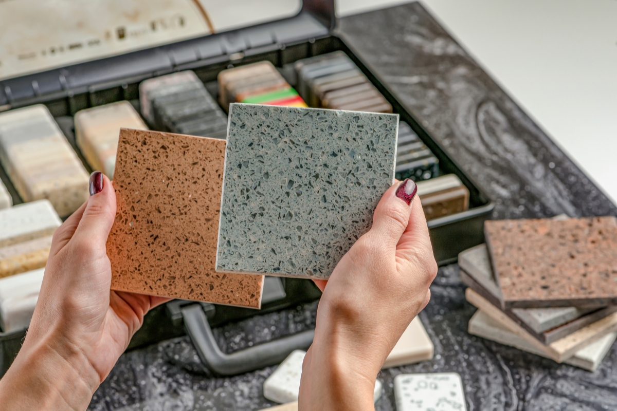 Elegant Bathroom Vanities in Roswell: Choosing the Right Style for Your Space 5 A person holds two countertop samples, one brown and one gray, over a table with more sample pieces and a display case of various material swatches—perfect choices for creating elegant bathroom vanities.