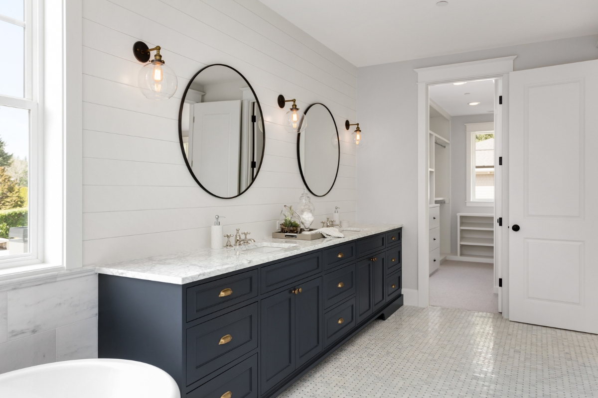 Elegant Bathroom Vanities in Roswell: Choosing the Right Style for Your Space 6 Modern bathroom with two round mirrors above elegant bathroom vanities in navy, brass fixtures, marble countertop, and white tiled floor; walk-in closet visible through open door.
