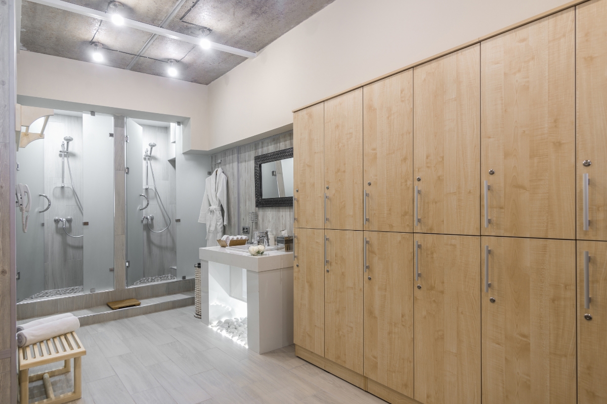 Elegant Bathroom Vanities in Roswell: Choosing the Right Style for Your Space 8 Modern locker room with wooden lockers, double shower area with glass dividers, elegant bathroom vanities stocked with towels and toiletries, and a bathrobe hanging near the showers.