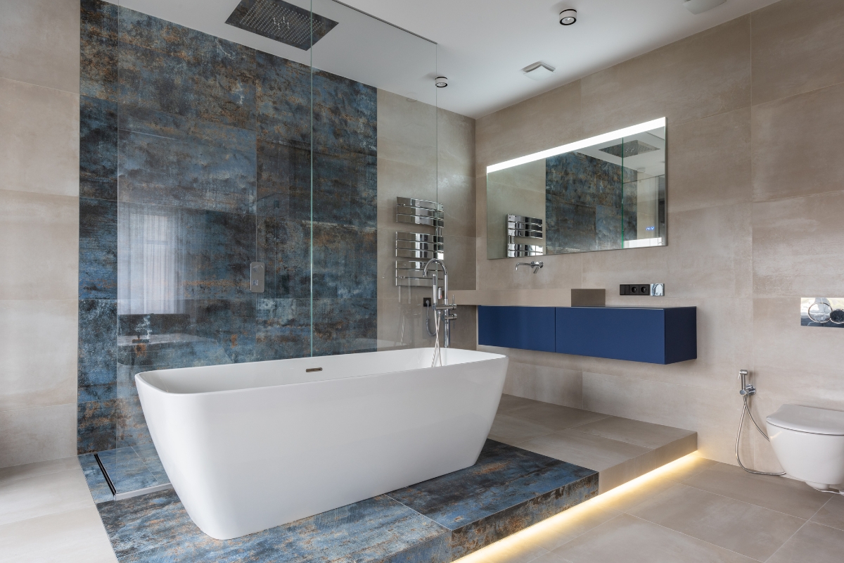 Modern bathroom with a freestanding white bathtub, blue accent wall and vanity, large mirror, wall-mounted toilet, bathroom tile flooring, and a glass-enclosed shower.
