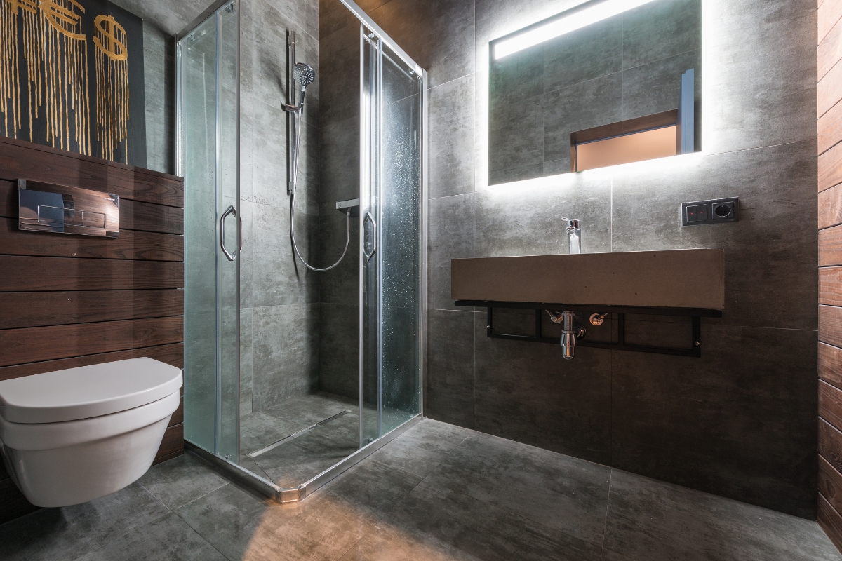 Bathroom Tile Flooring in Vinings: Expert Tips, Trends, and Benefits 6 Modern bathroom with glass shower enclosure, wall-mounted toilet, rectangular sink, large backlit mirror, and sleek bathroom tile flooring on the gray walls and floor.