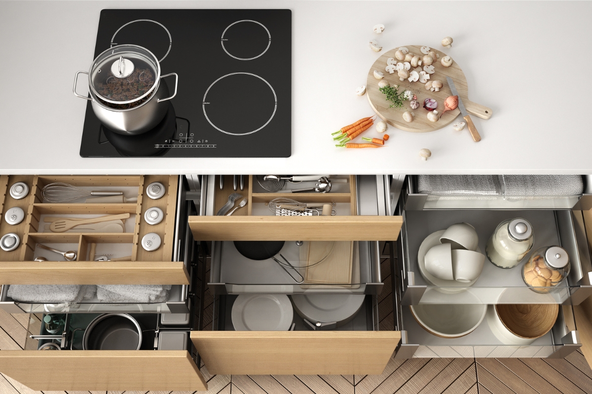 Custom Kitchen Storage Solutions in Kennesaw: Maximize Your Space 2 A kitchen countertop with a cooktop, pot, and cutting board with vegetables sits above open drawers featuring custom kitchen storage for utensils, plates, bowls, jars, and other kitchenware.