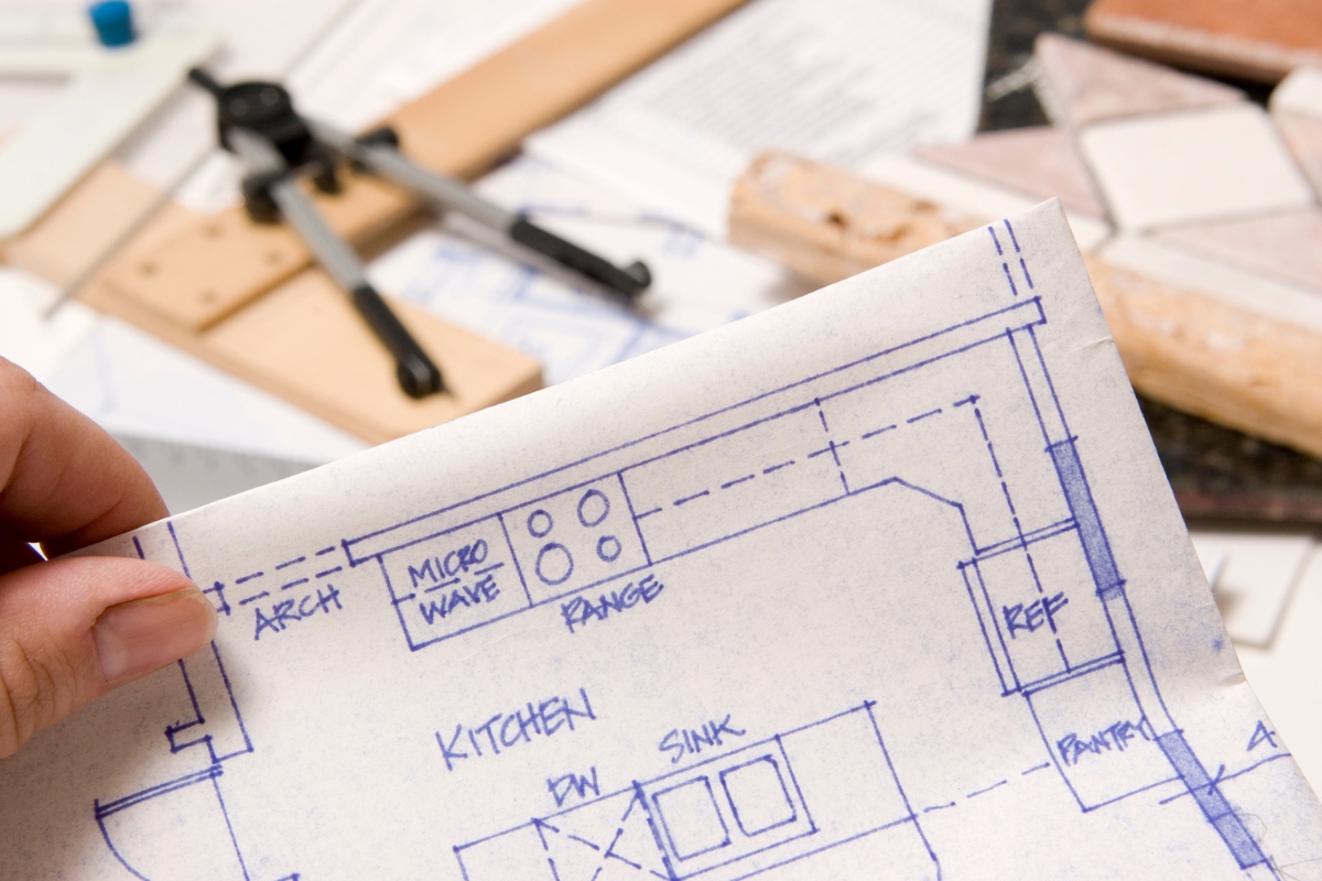 Eco-Friendly Kitchen Remodeling in Smyrna: Sustainable Upgrades That Last 16 A hand holds a kitchen blueprint with notes, while drafting tools, paperwork, and tile samples for an eco-friendly kitchen remodeling project are scattered on a work surface in the background.