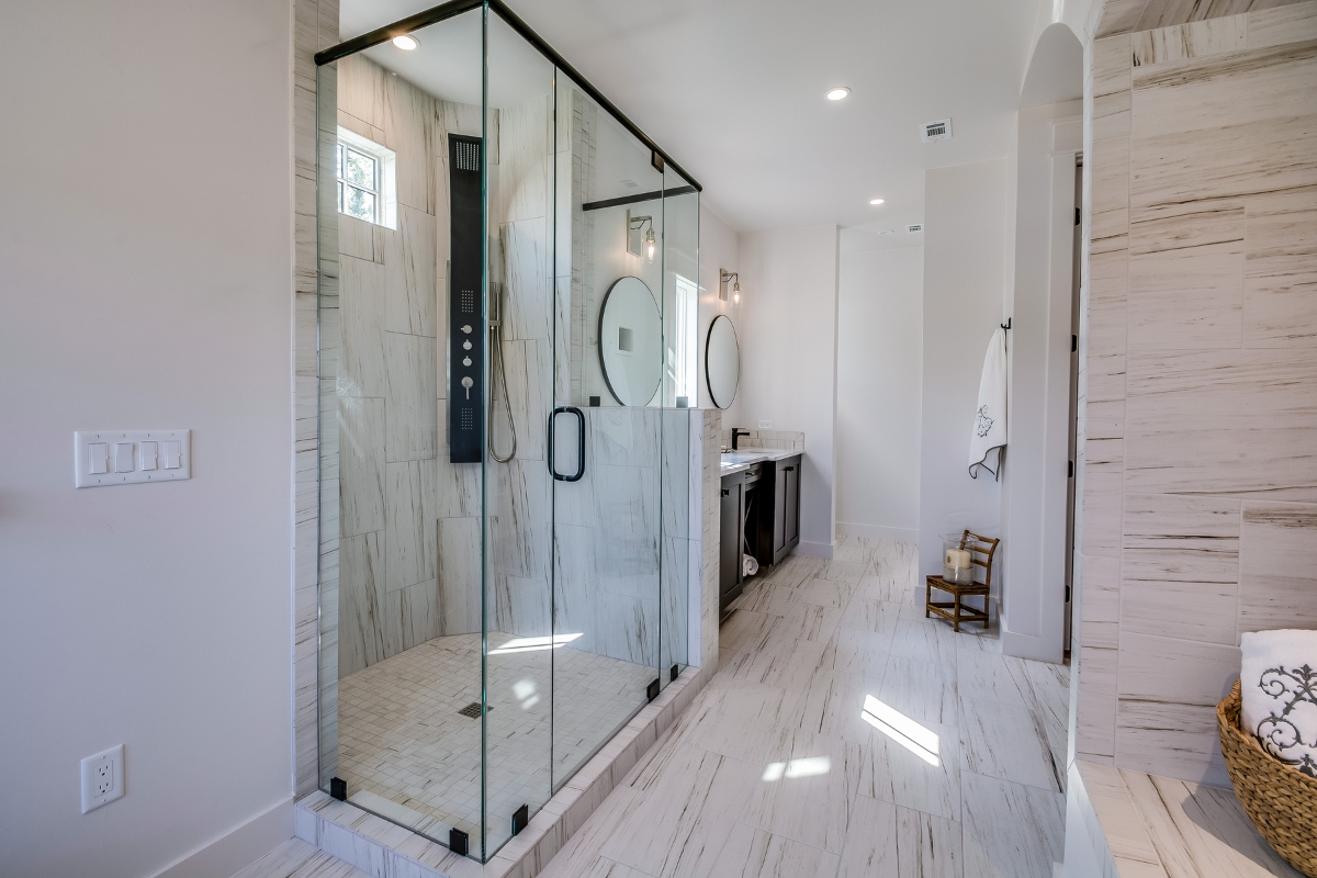 Shower Replacement in Sandy Plains: What to Expect and How to Prepare 5 Modern bathroom with a large glass-enclosed shower replacement, dual vanities with round mirrors, white tile flooring, and a towel basket on the right.