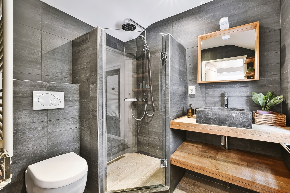 Modern bathroom with dark gray tiles, stylish shower replacement featuring a glass enclosure, wall-mounted toilet, wooden vanity with black basin, mirror, and a small potted plant.