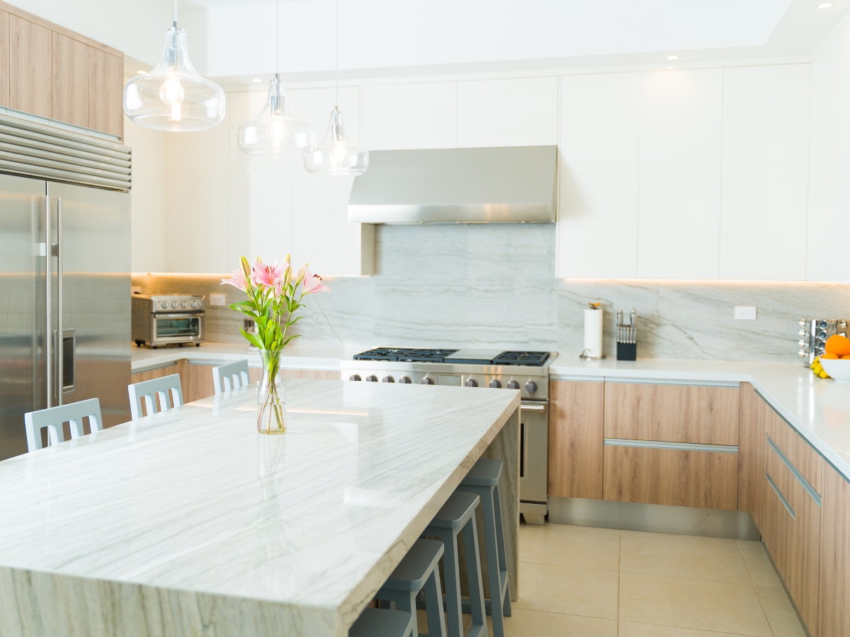 Quartz vs. Granite Countertops in Alpharetta: Which Should You Choose? 11 Modern kitchen with a marble island, wooden cabinets, stainless steel appliances, pendant lights, and a vase with pink flowers on the countertop—perfect for exploring Quartz vs. Granite Countertops in style and elegance.