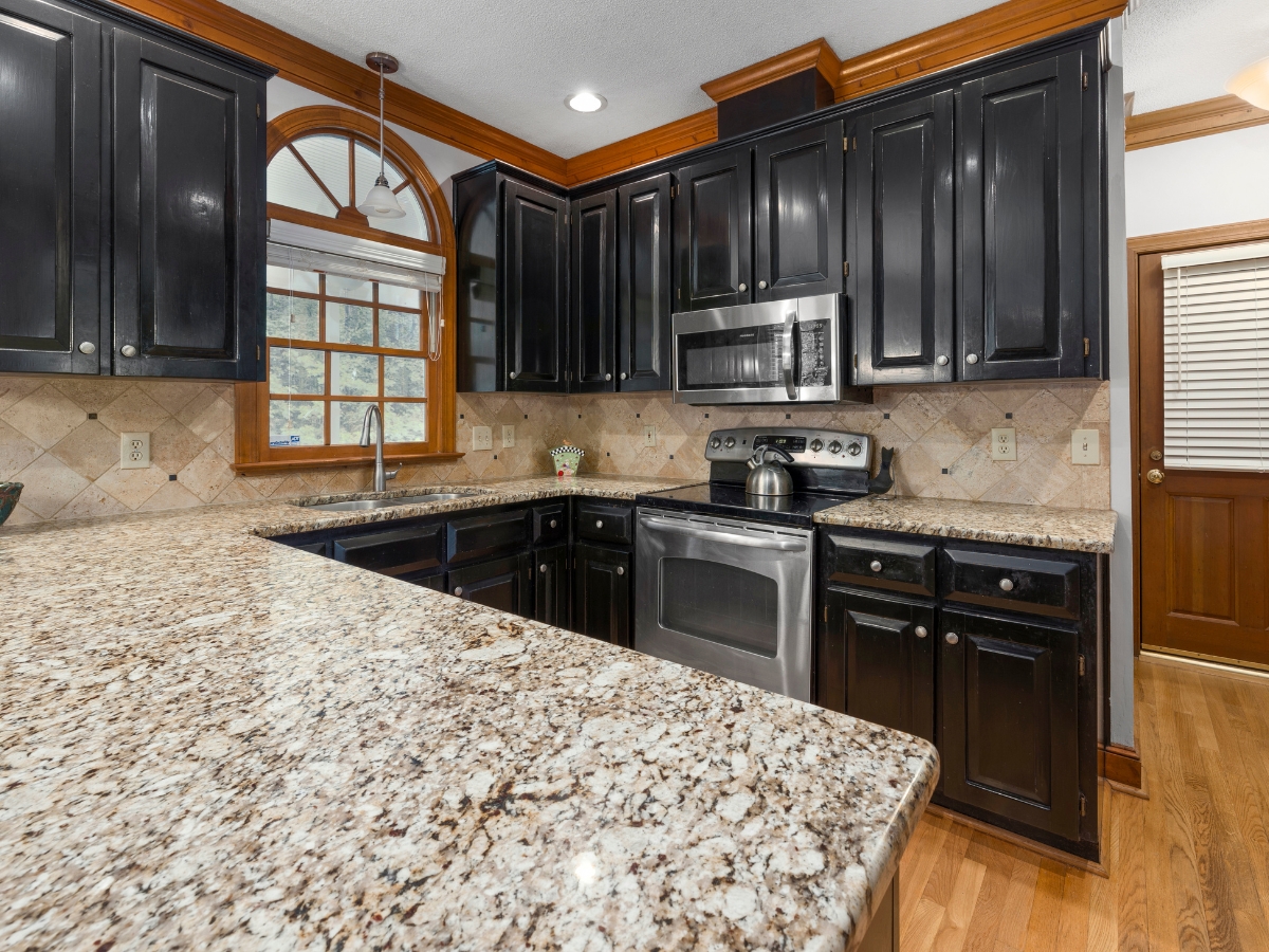 Quartz vs. Granite Countertops in Alpharetta: Which Should You Choose? 13 Modern kitchen with black cabinets, stainless steel appliances, and granite countertops—explore the Quartz vs. Granite Countertops debate in a space featuring a tile backsplash, hardwood floors, large window over the sink, and a wooden door in the background.