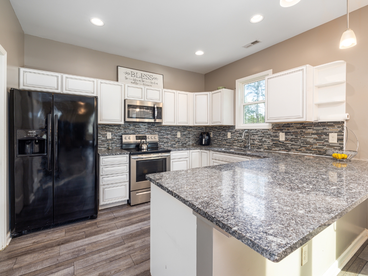 Quartz vs. Granite Countertops in Alpharetta: Which Should You Choose? 15 Modern kitchen with white cabinets, black appliances, gray tile backsplash, and wood flooring. Granite countertops add elegance—perfect for those comparing Quartz vs. Granite Countertops. A window sits above the sink and a decorative sign hangs above the cabinets.