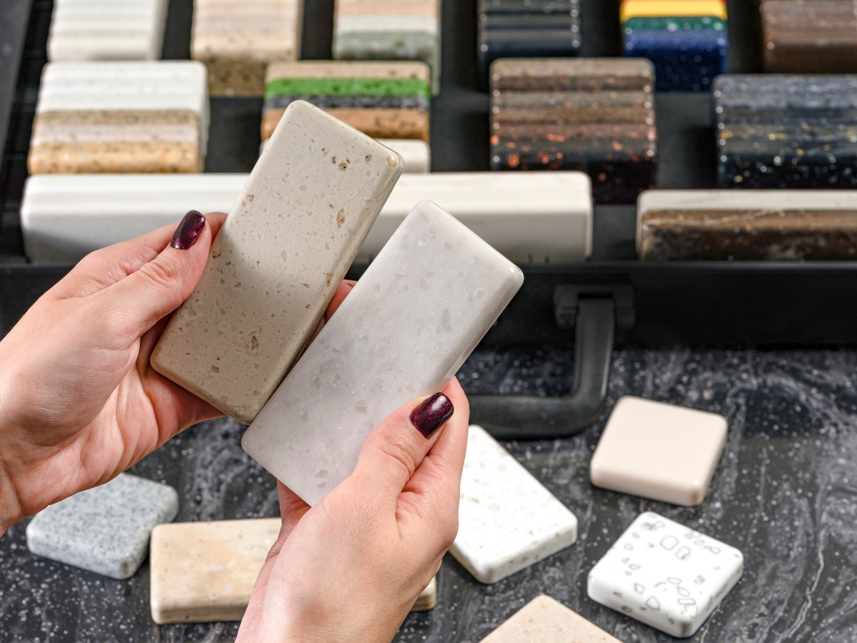 Quartz vs. Granite Countertops in Alpharetta: Which Should You Choose? 9 A person holds two stone countertop samples over a table with various others, comparing Quartz vs. Granite Countertops, with a case containing even more stone pieces nearby.
