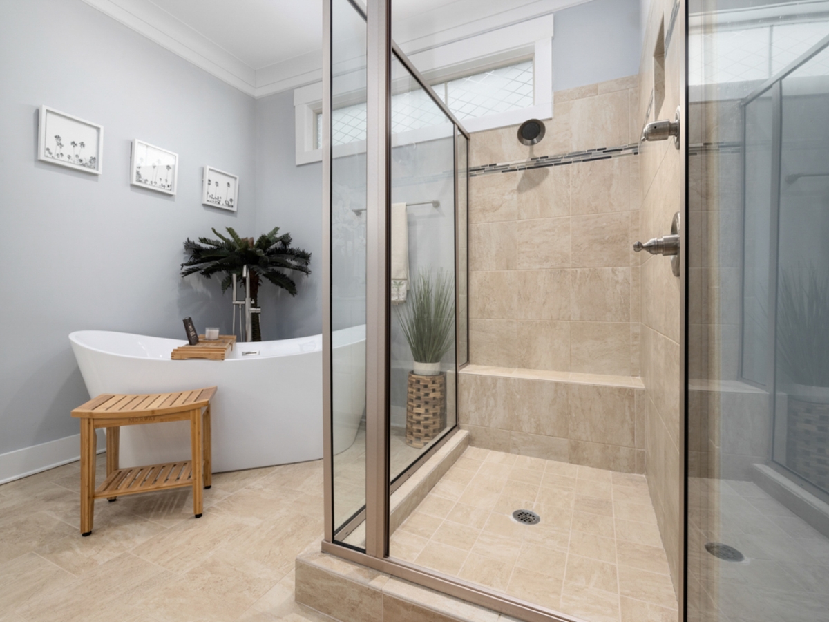 Walk-In Showers vs. Bathtubs in Johns Creek: Which Is Right for You? 11 Modern bathroom with a glass-enclosed walk-in shower, beige tile, a white freestanding bathtub, a wooden stool, and decorative plants.