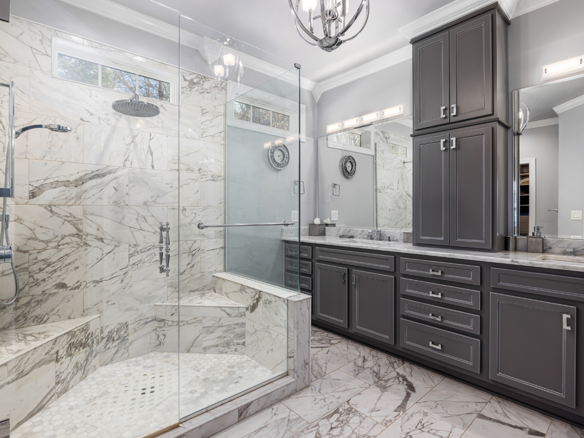 Walk-In Showers vs. Bathtubs in Johns Creek: Which Is Right for You? 14 Modern bathroom with marble tile, glass shower enclosure with a bench, rainfall showerhead, and gray cabinetry featuring double sinks and mirrors—perfect for those who love stylish walk-in showers.