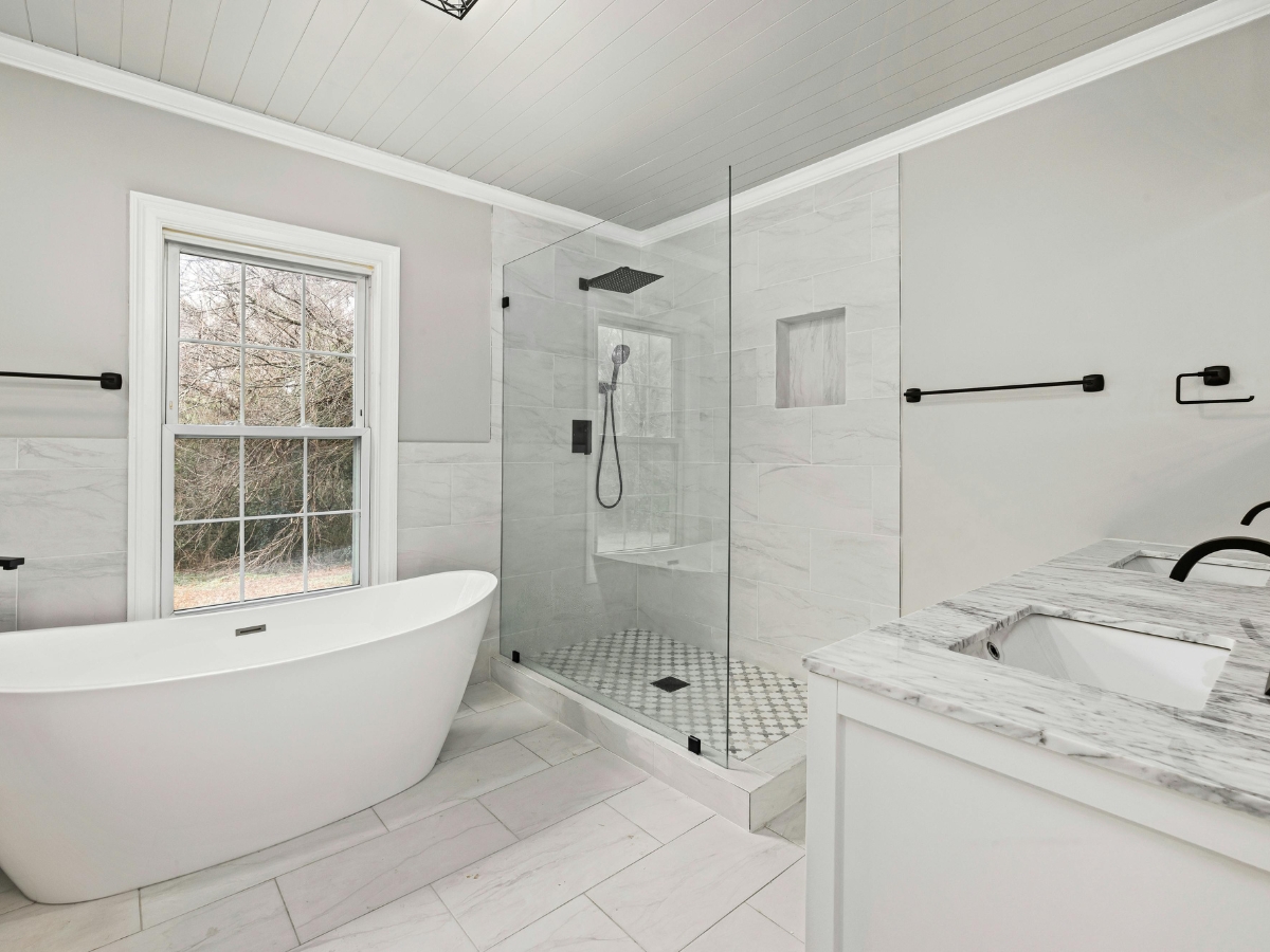 Walk-In Showers vs. Bathtubs in Johns Creek: Which Is Right for You? 15 Modern bathroom with a freestanding bathtub, elegant walk-in showers, marble countertop vanity, and a large window letting in natural light.