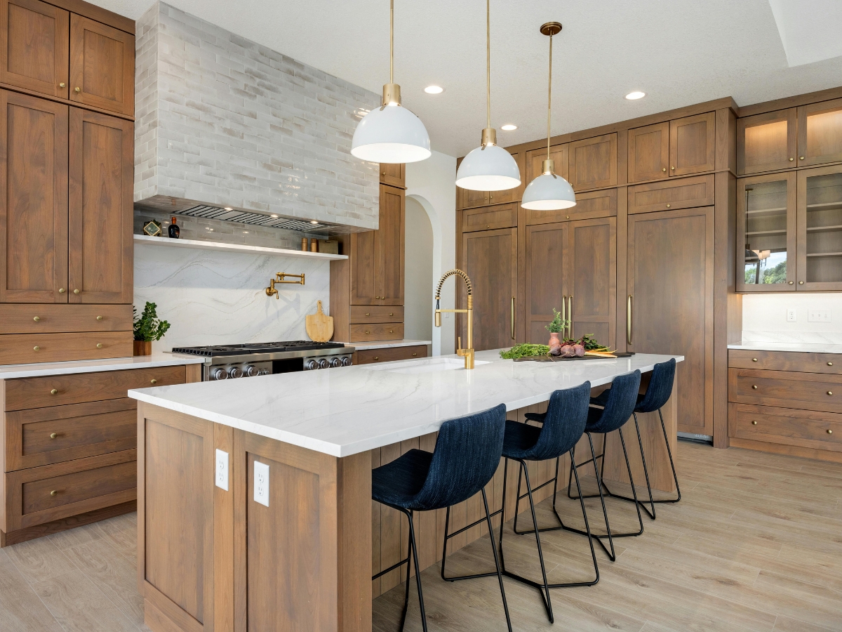 Custom Cabinetry vs. Stock Cabinets in Sandy Springs: Which Is Right for You? 3 Modern kitchen with wood cabinets, a large white marble island, gold fixtures, pendant lights, and four dark blue barstools—showcasing the beauty of custom cabinetry vs. stock cabinets. Stainless steel appliances and decorative plants complete the look.