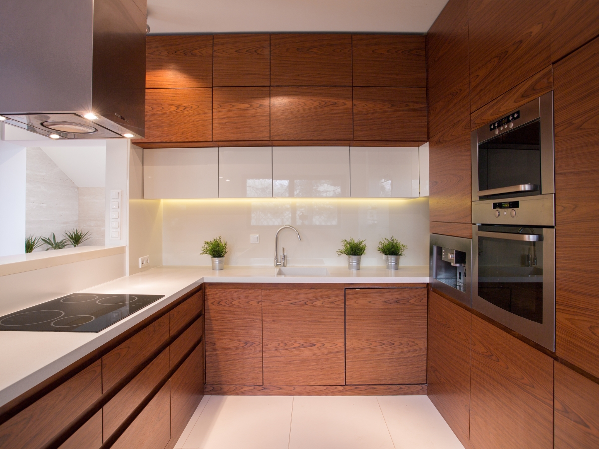 Custom Cabinetry vs. Stock Cabinets in Sandy Springs: Which Is Right for You? 2 Modern kitchen with custom cabinetry vs. stock cabinets, built-in ovens, electric cooktop, white countertops, under-cabinet lighting, and small potted plants by the sink.