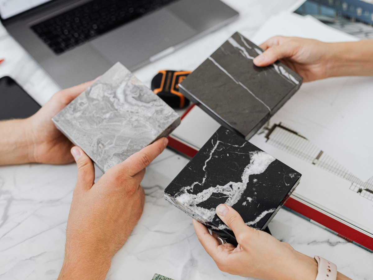 Transform Your Bathroom with a Modern Bathroom Vanity in Buckhead GA 4 Three hands hold different marble samples—one gray, one dark gray, and one black with white veins—over a marble table next to a laptop and architectural plans, considering options for a modern bathroom vanity.