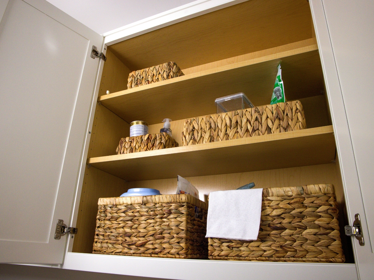 Essential Tips for Organizing Kitchen Cabinets 4 Open white cabinet with three shelves holding woven baskets and assorted toiletries, such as a hand towel, lotion, and a pump bottle—showcasing clever tips for organizing kitchen cabinets or bathroom essentials neatly.
