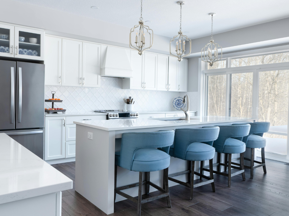 Modern kitchen with white cabinets, stainless steel appliances, a large island, and four blue upholstered bar stools under pendant lights. Large windows provide natural light—perfect for those seeking budget kitchen staging ideas.