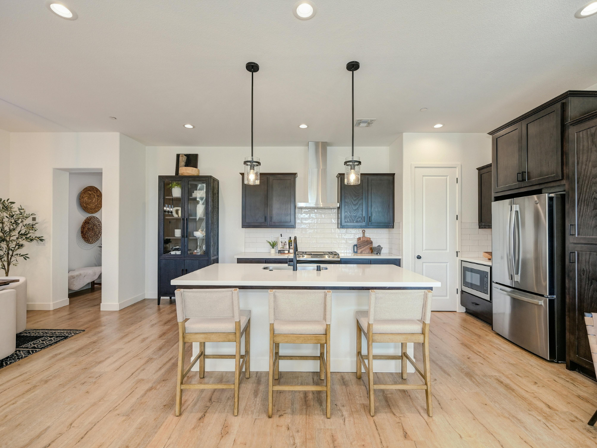 Modern kitchen with a central island, three chairs, pendant lights, stainless steel appliances, dark cabinets, and light wood flooring—perfect for those seeking budget kitchen staging ideas.