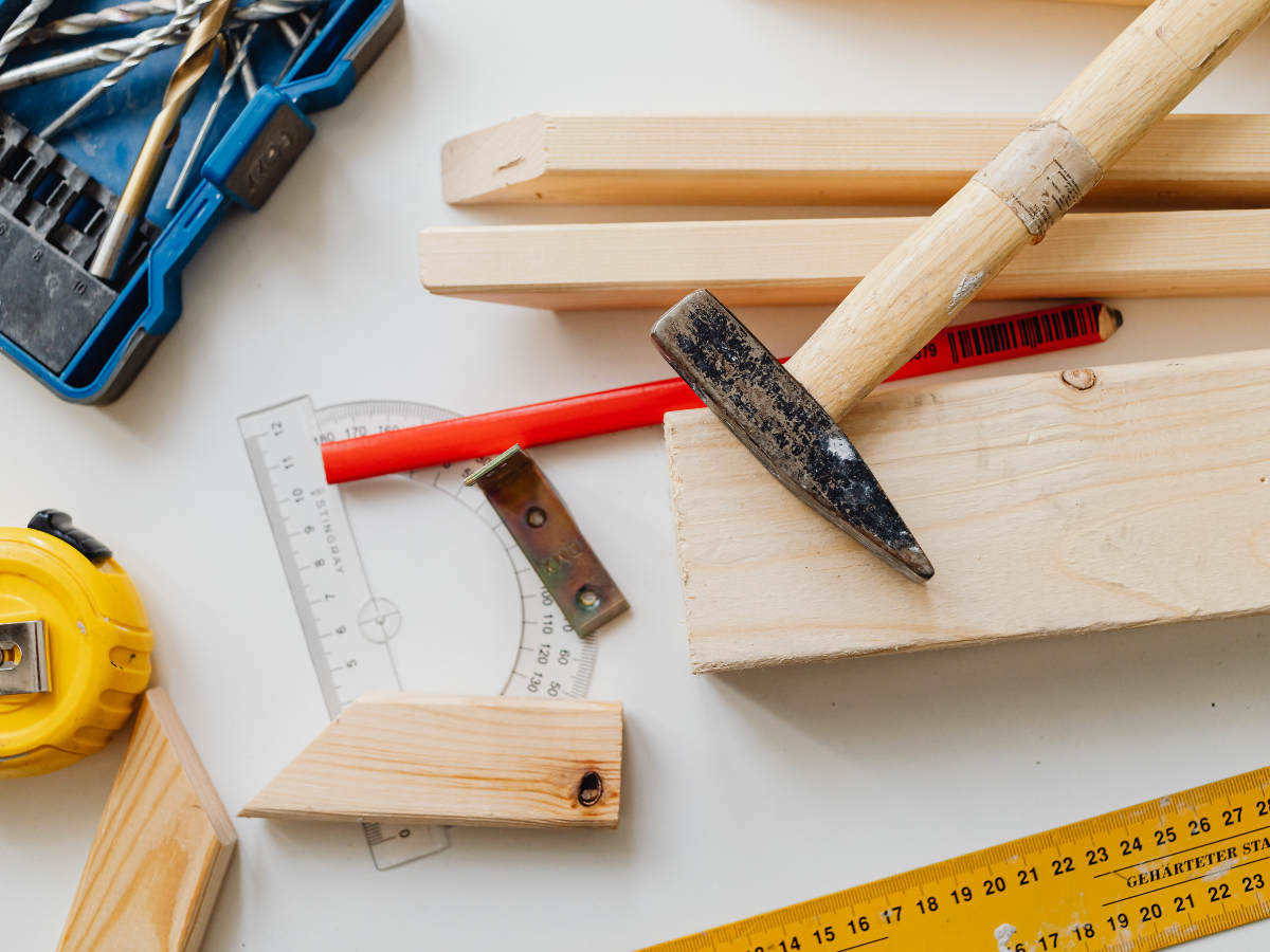 Woodworking tools and materials, perfect for budget kitchen staging ideas, including a hammer, wooden planks, protractor, pencil, tape measure, and drill bits, are arranged on a white work surface.