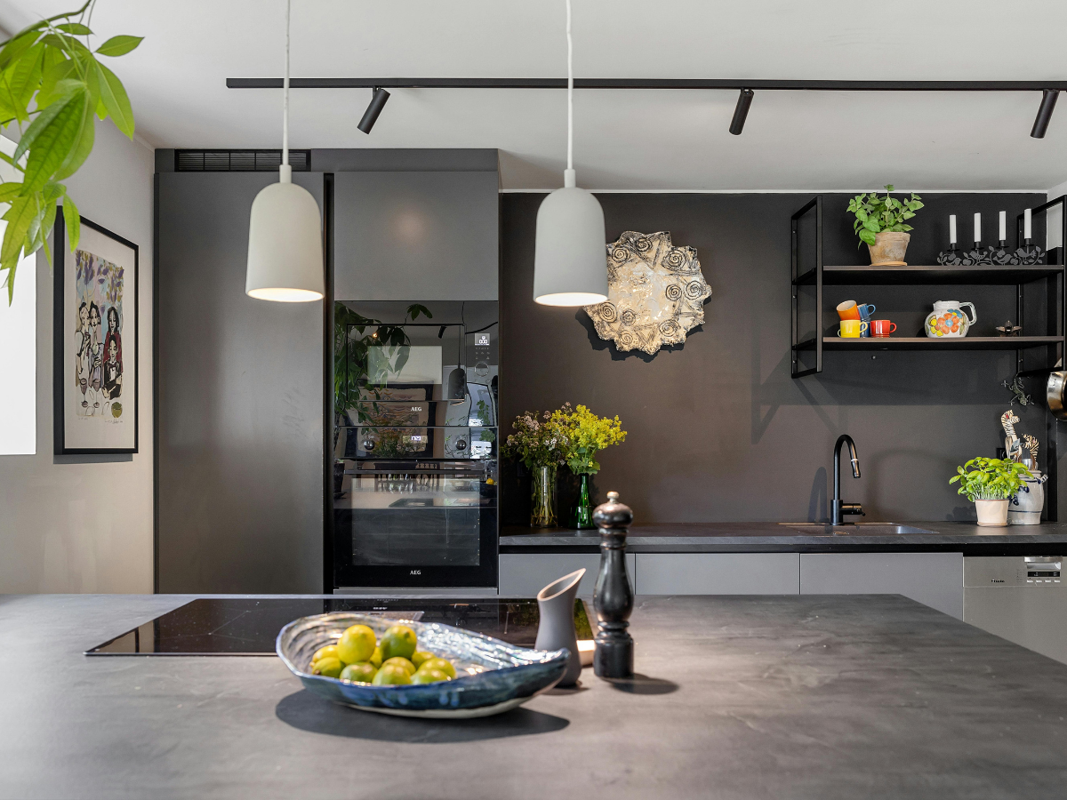 Modern kitchen with dark cabinets, a black countertop, hanging pendant lights, wall art, potted plants, and a bowl of green fruit on an island—showcasing smart budget kitchen staging ideas.