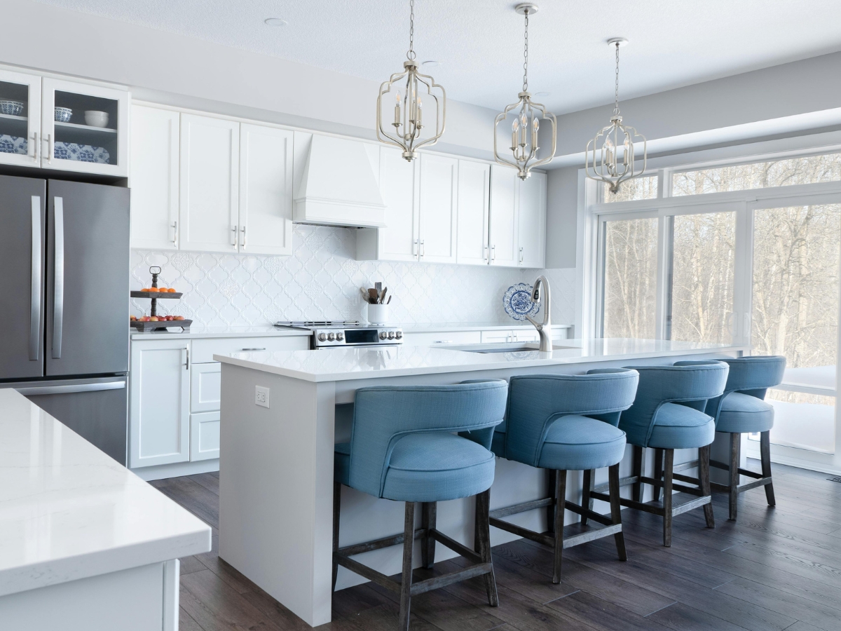 Modern kitchen with white cabinets, stainless steel appliances, a large island, and four blue upholstered barstools. Avoid common kitchen staging mistakes to let the three pendant lights and expansive windows truly enhance the space.