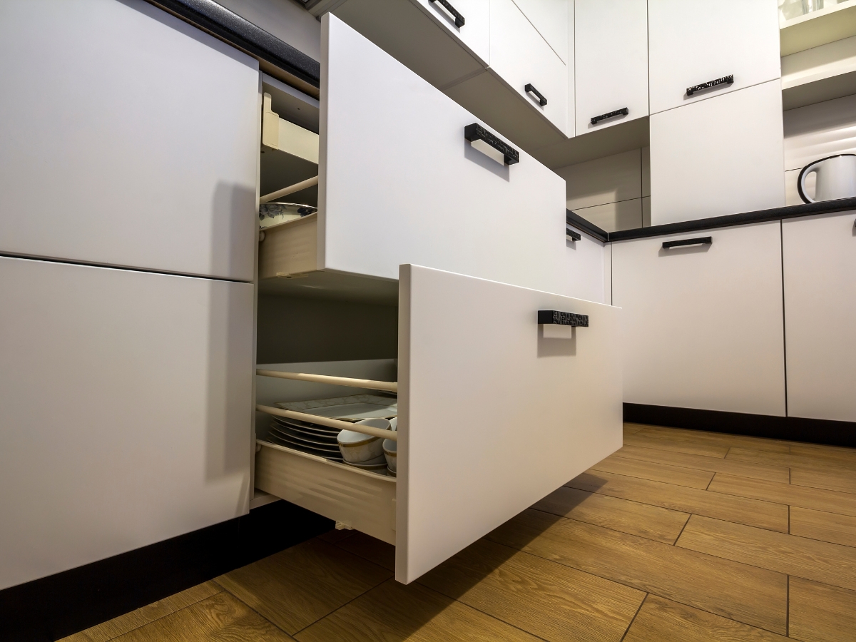 White kitchen drawers are open, revealing plates and cups inside. The floor is wooden, and the cabinets have black handles—a setup that avoids common kitchen staging mistakes by keeping items neat and surfaces clutter-free.
