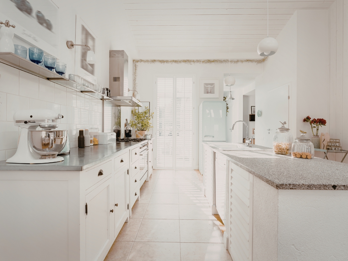 Bright, modern kitchen with white cabinets, appliances, and countertops; sunlight streams through French doors at the far end, creating an airy atmosphere free from common kitchen staging mistakes.