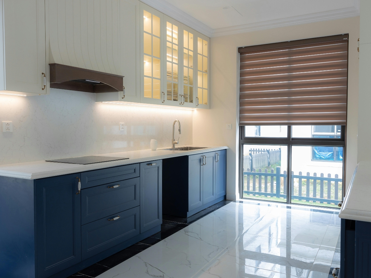 Modern kitchen with blue lower cabinets, white countertops, glass-front upper cabinets, induction cooktop, sink, and a large window with brown blinds—perfect inspiration to choose kitchen colors that create a fresh and inviting space.