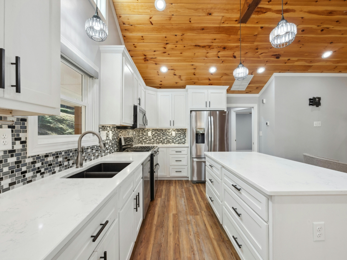 Modern kitchen staging with white cabinets, black hardware, stainless steel appliances, mosaic tile backsplash, wood ceiling, pendant lights, and a large center island with white countertops.