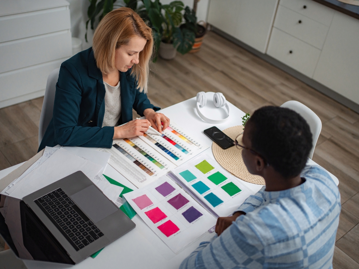 Two people sit at a table with fabric swatches and color samples, discussing Kitchen Staging choices. A laptop, headphones, and documents are on the table.