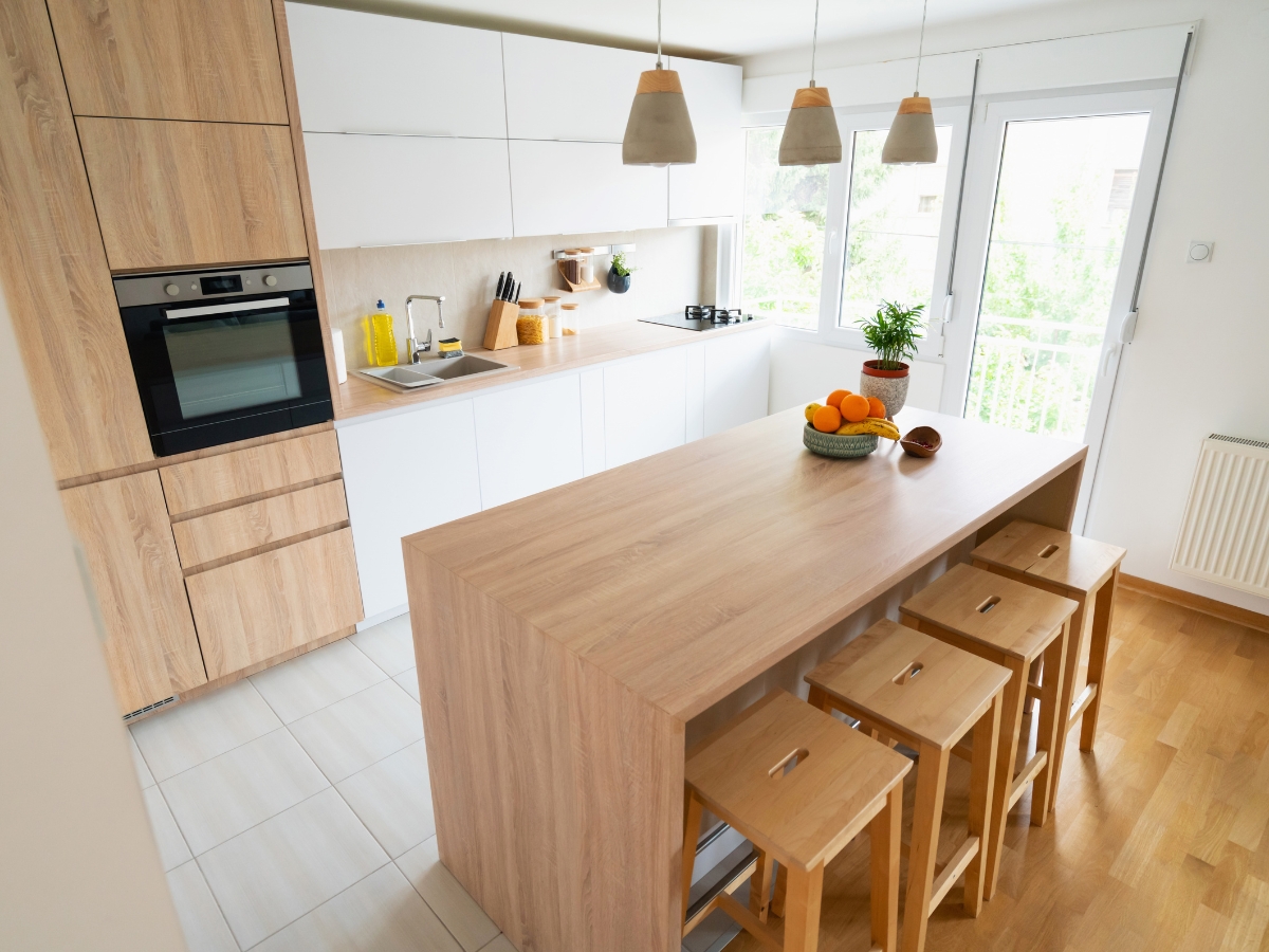 Modern kitchen staging with light wood cabinets, white countertops, a built-in oven, island with four stools, pendant lights, and a bowl of fruit on the island.