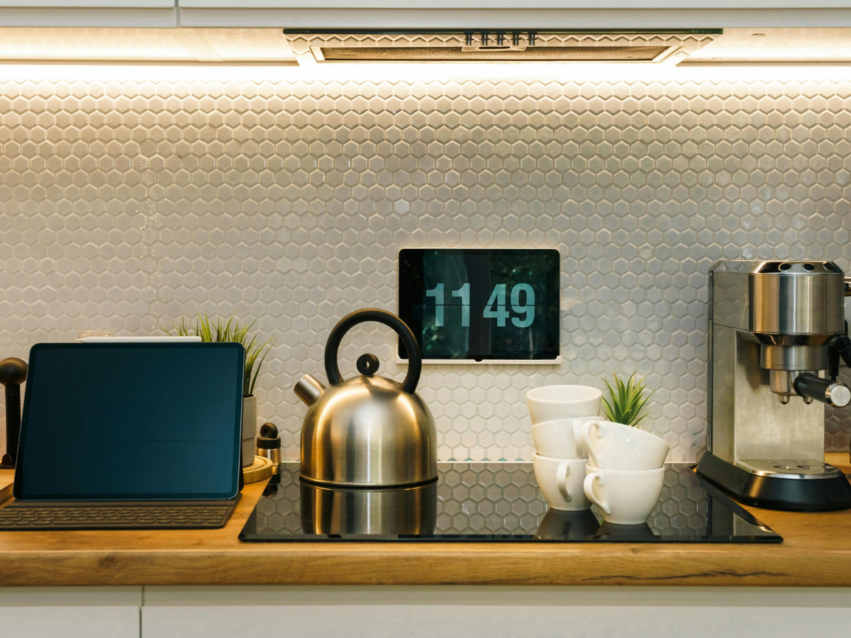 A modern kitchen counter with energy saving appliances, a laptop, a stainless steel kettle, stacked white cups, a coffee machine, small plants, and a digital clock showing 11:49 on the backsplash.