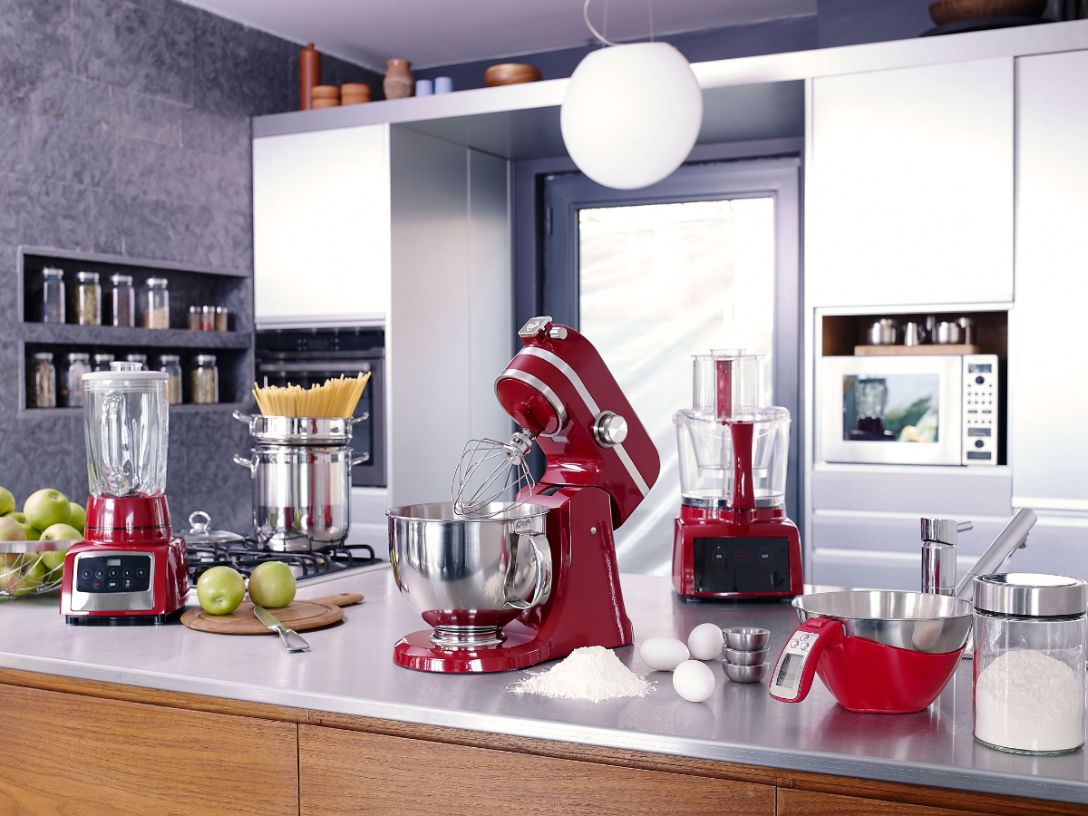 Modern kitchen with red, energy saving appliances—including a blender, stand mixer, and food processor—on a countertop with baking ingredients like eggs, flour, and apples.