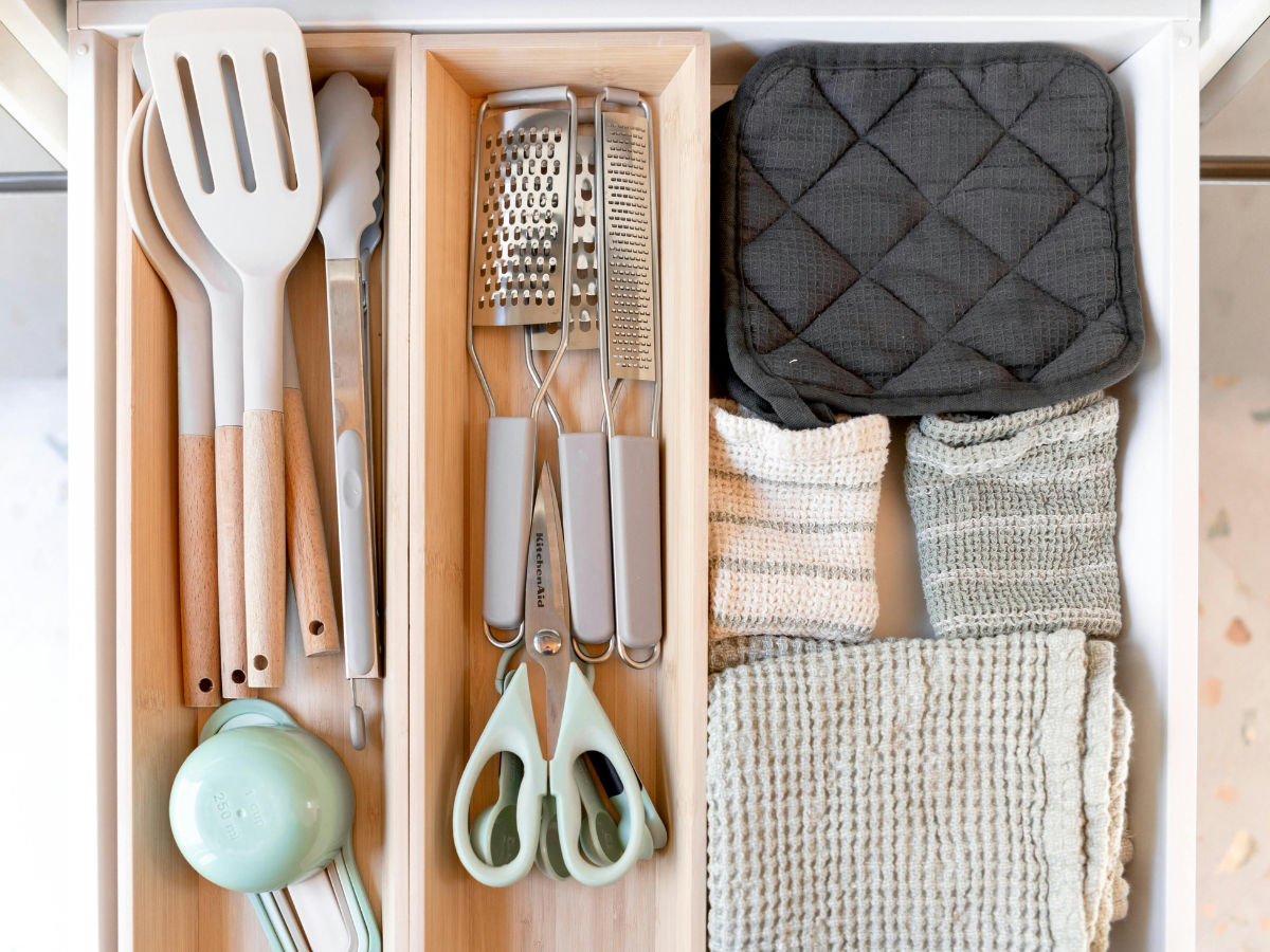 Maximize Space in Your Small Kitchen Today: Expert Storage Ideas and Efficient Layouts 5 Kitchen drawer with utensils, metal tongs, graters, scissors, measuring cups, and folded dish towels and oven mitt arranged in wooden organizers to help maximize space in your small kitchen today.