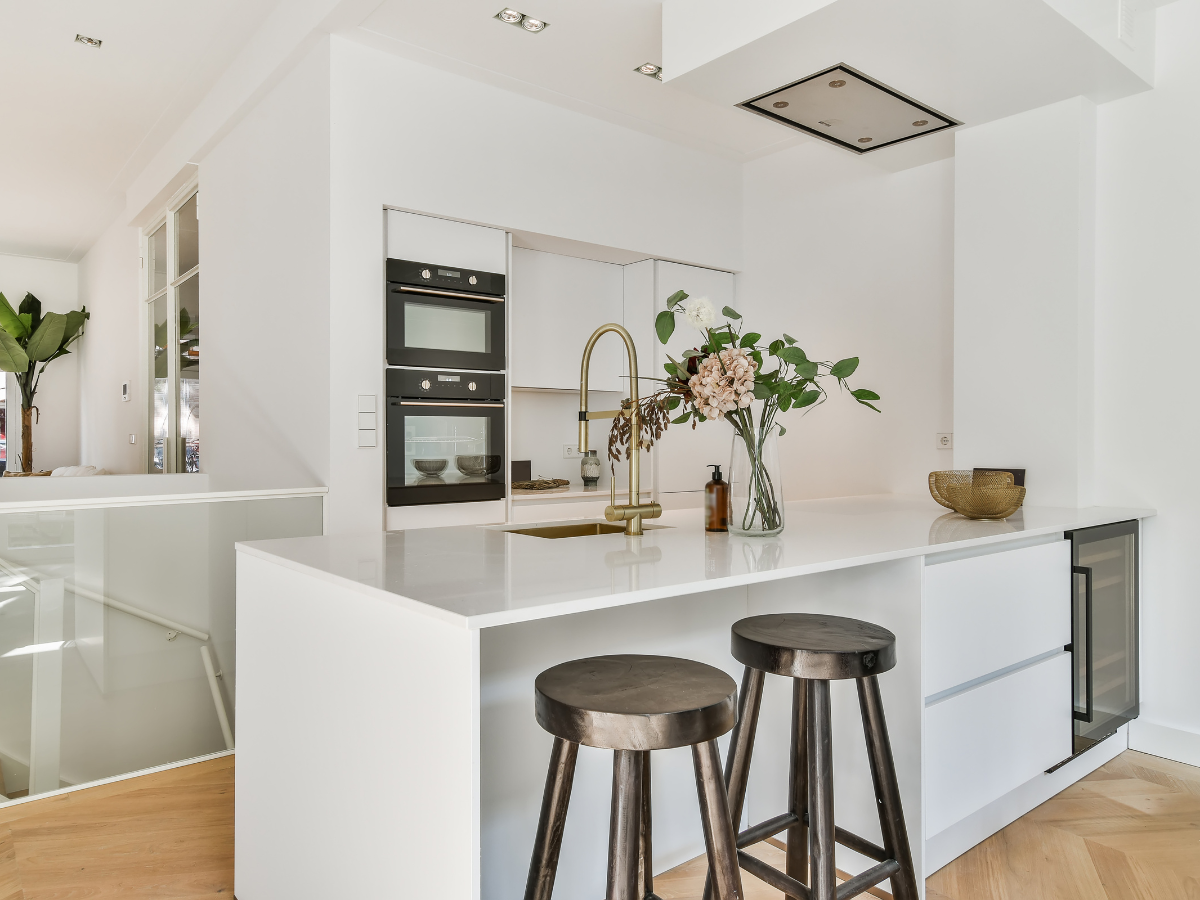 Kitchen Staging Ideas to Wow Potential Buyers: Expert Tips for Effective Home Selling 5 Modern white kitchen with a central island, two bar stools, a gold faucet, built-in ovens, and a vase of flowers on the counter—perfect staging ideas for showcasing style and functionality.