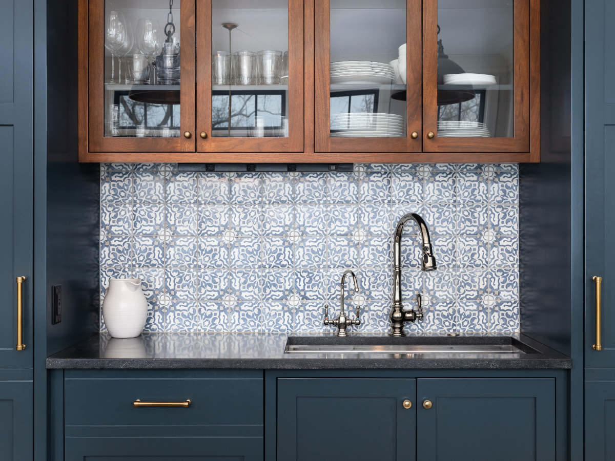 Kitchen sink area with a patterned blue and white affordable backsplash tile, blue cabinets, brass hardware, glass-front upper cabinets, and a white pitcher on the counter.