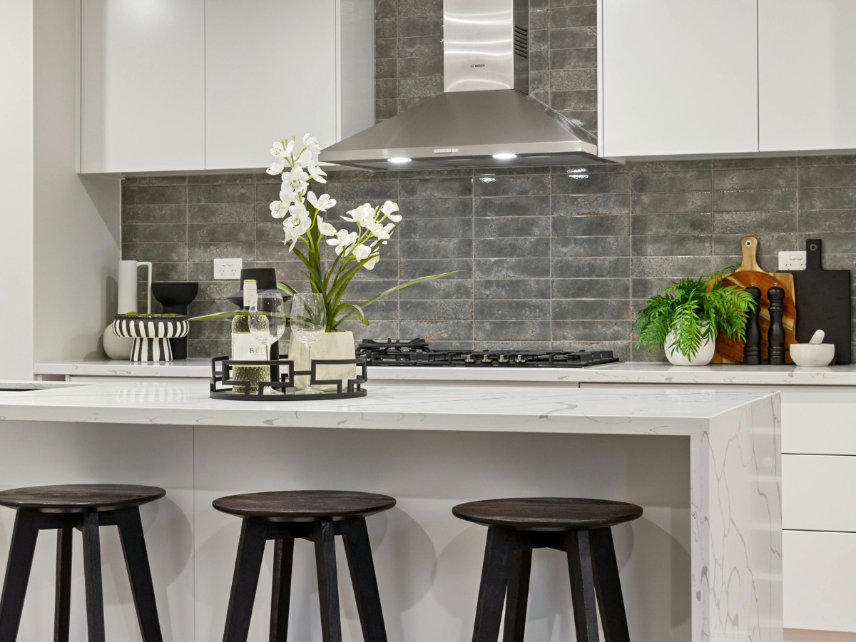 Modern kitchen with white cabinets, marble island, three dark stools, a cooktop with a range hood, and decorative plants and accessories on the counter and affordable backsplash tile.