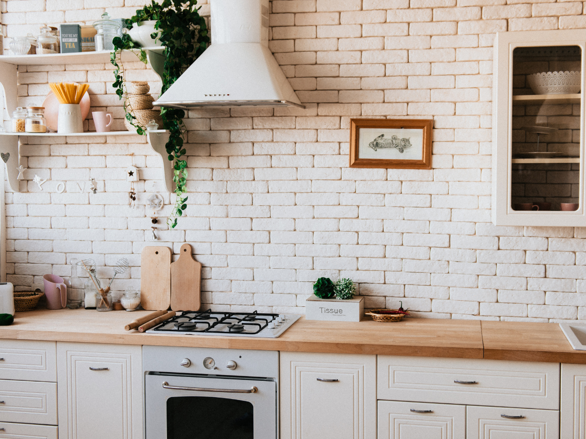 Modern kitchen with white cabinets, affordable backsplash tile in a stylish brick pattern, open shelves holding dishes and plants, a stove with oven, a range hood, and framed artwork on the wall.