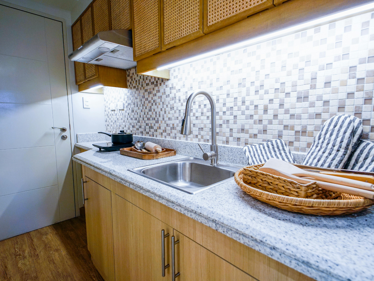 Modern kitchen with light wood cabinets, a stainless steel sink, electric stove, affordable backsplash tile in a mosaic pattern, and utensils in a wicker basket on the countertop.