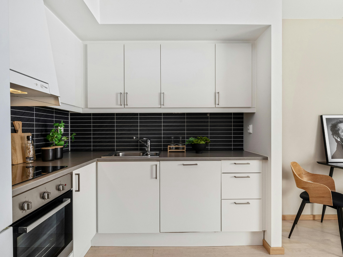 Modern integrated kitchen with white cabinets, black tile backsplash, stainless steel oven, sink, and small plants; adjacent dining area visible with a wooden chair and framed photo on the wall.