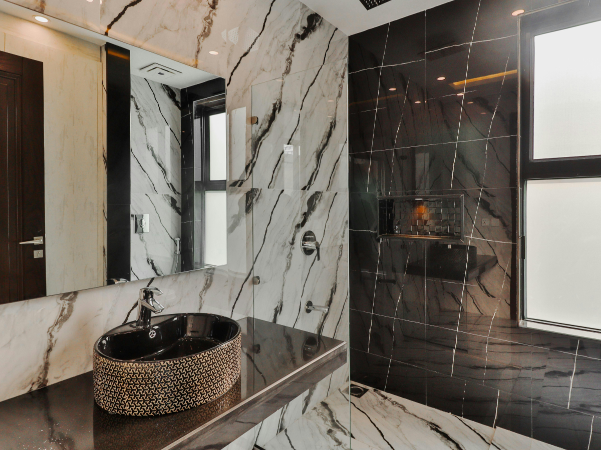 Modern bathroom with marble-patterned walls, a large mirror, a circular black sink with a patterned exterior, and a window allowing natural light—perfectly complementing an integrated kitchen in a contemporary home.